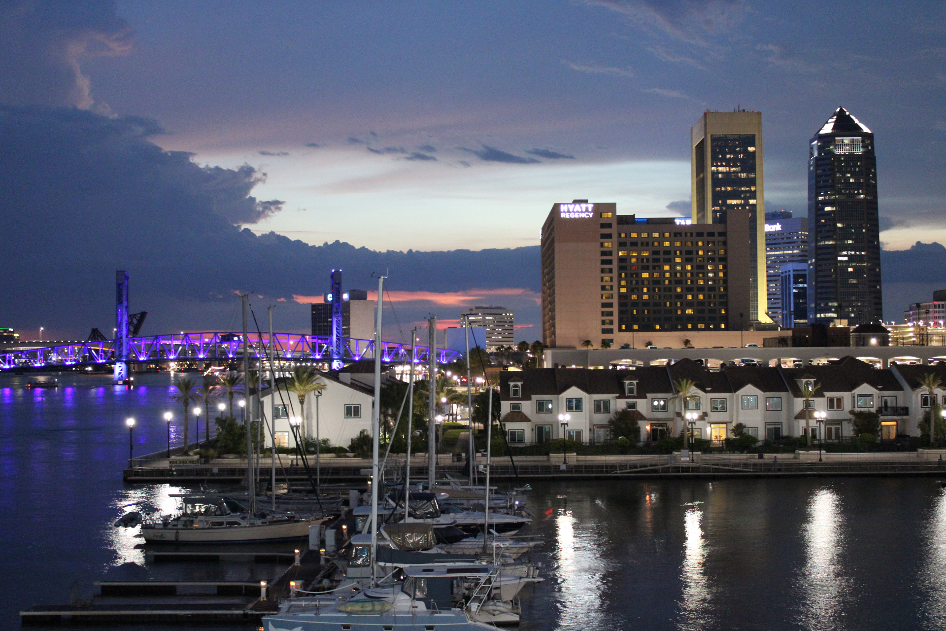 Jacksonville downtown skyline at dusk with the Main Street Bridge lit purple, the Hyatt Regency, and sailboats in the marina