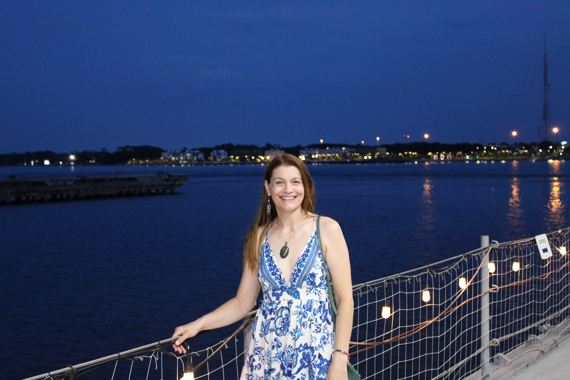 Guest in a blue floral dress smiling on the pier beside the USS Orleck at night with the Jacksonville riverfront lights across the St. Johns River